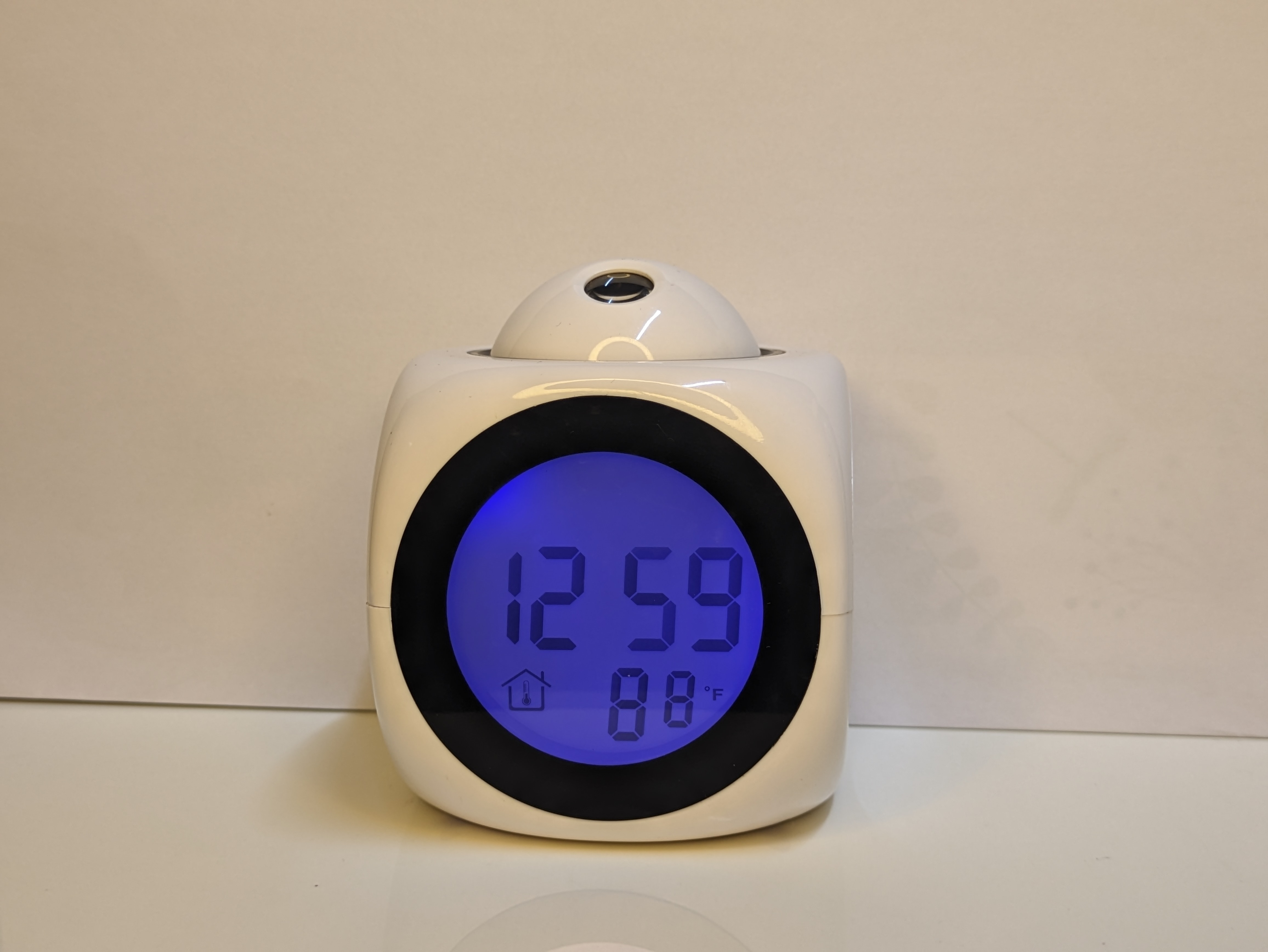 A rounded cube-shaped digital talking table clock image, with a led screen displaying the time on its body as 12:59, with a round protruding pressable top, 